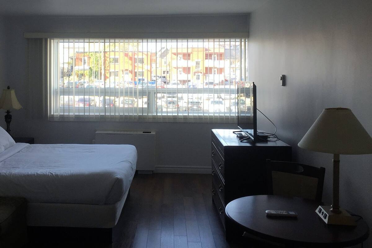 A hotel room with a bed, a desk, a chair, a lamp, and a window with a view of buildings and parked cars.