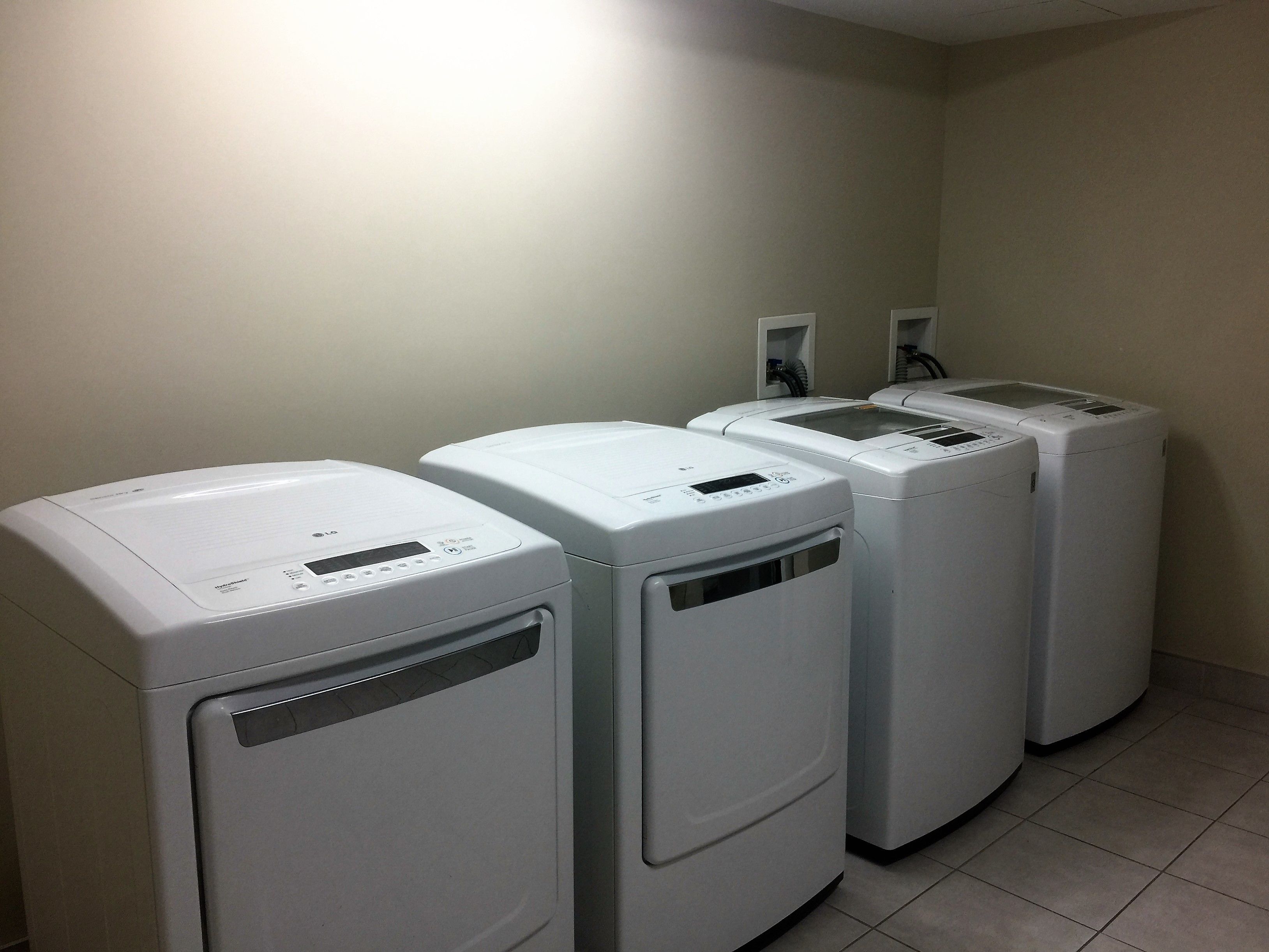 A row of four white laundry machines in a room with plain walls and tiled flooring.