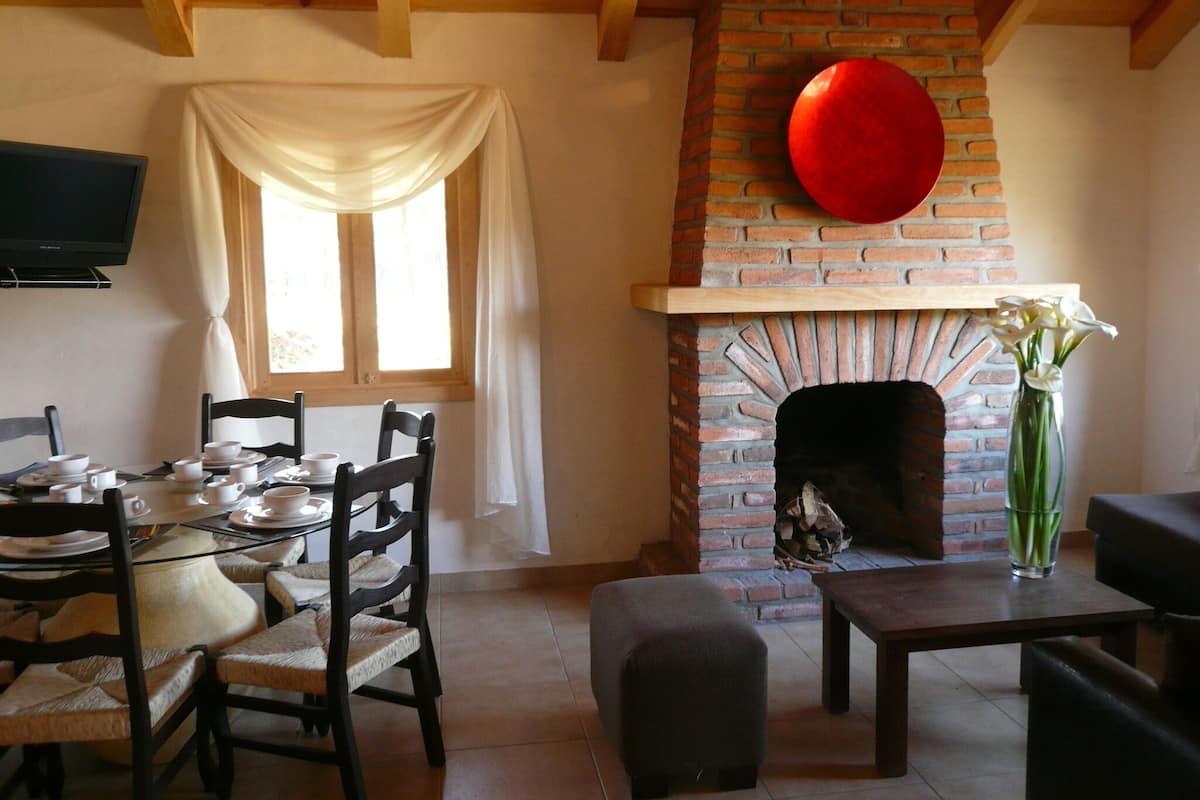 A cozy living room with a brick fireplace, wooden beams, a dining area with chairs and a table set for a meal, and a television mounted on the wall.