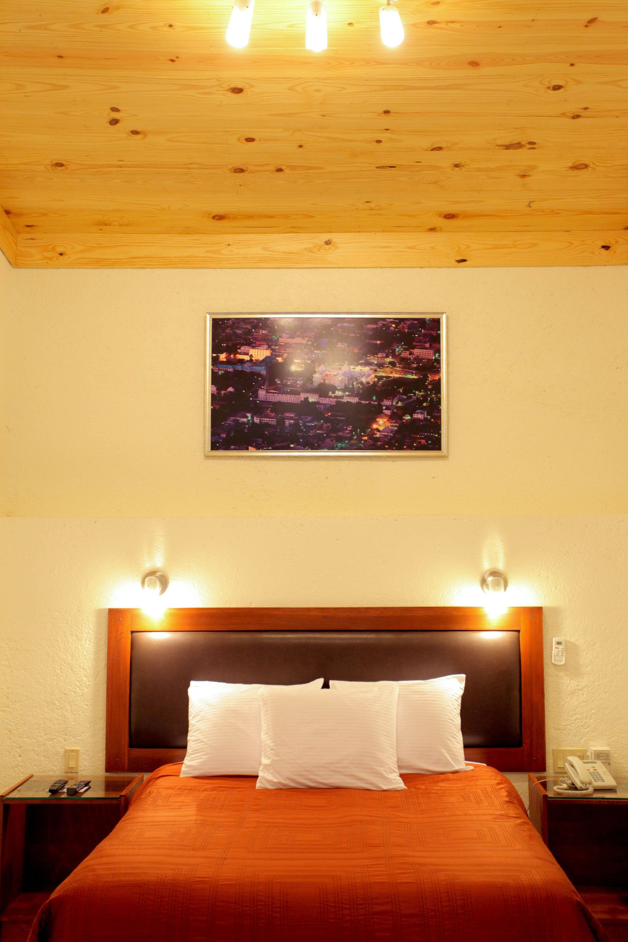 A hotel room with a wooden ceiling, a cityscape picture on the wall, a bed with white pillows, and bedside lamps.