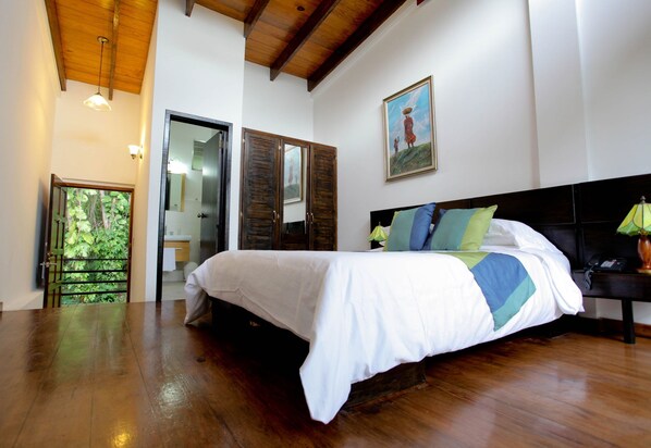 A bedroom with a wooden floor, a bed with a white and blue bedspread, a wooden headboard, and a painting on the wall.