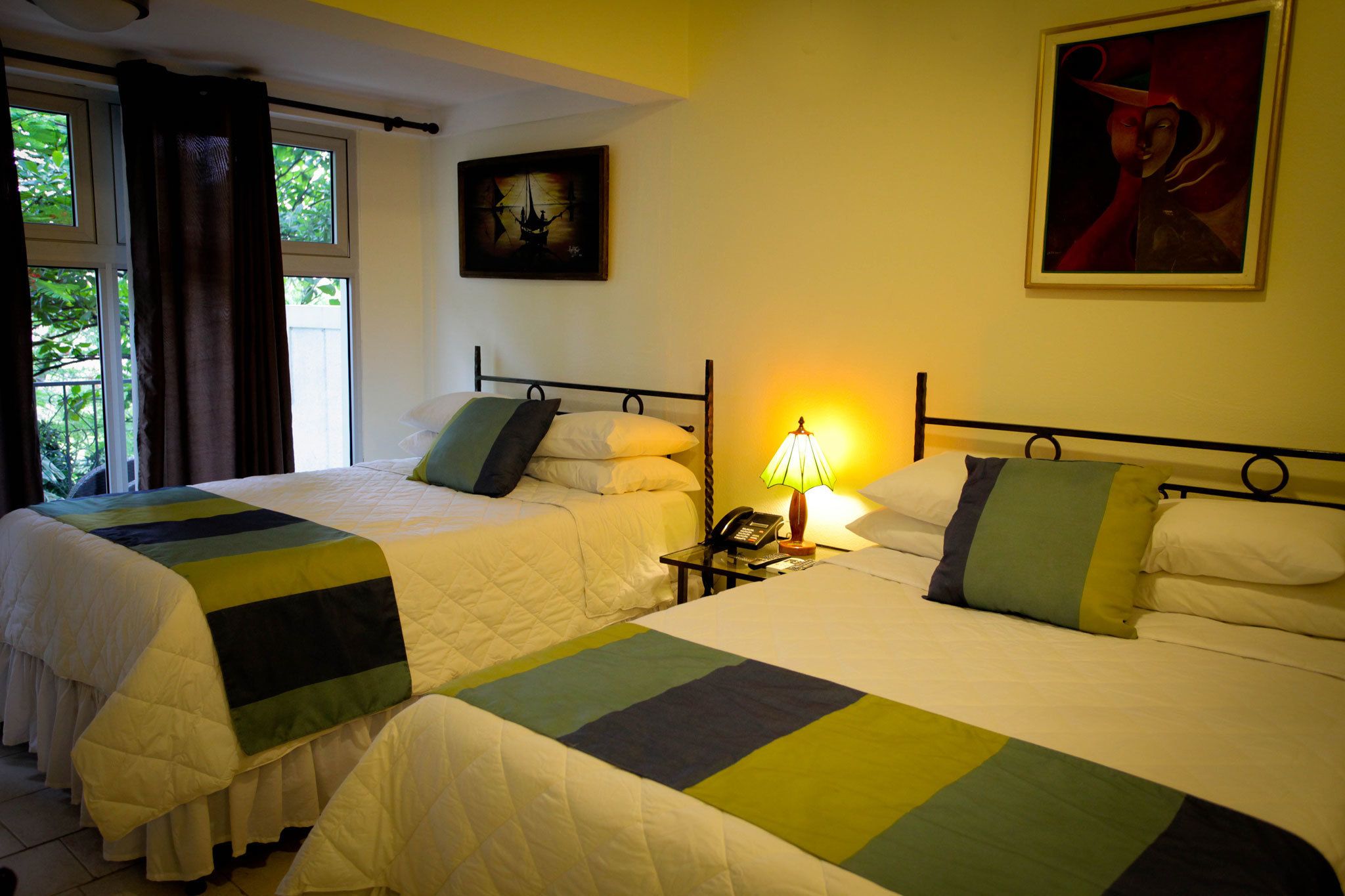 A hotel room with two beds, a bedside table with a lamp, a painting on the wall, and a window with curtains.