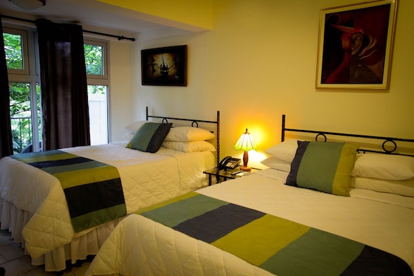 A hotel room with two beds, a bedside table with a lamp, a painting on the wall, and a window with curtains. - La Reserve Hotel (Petionville)
