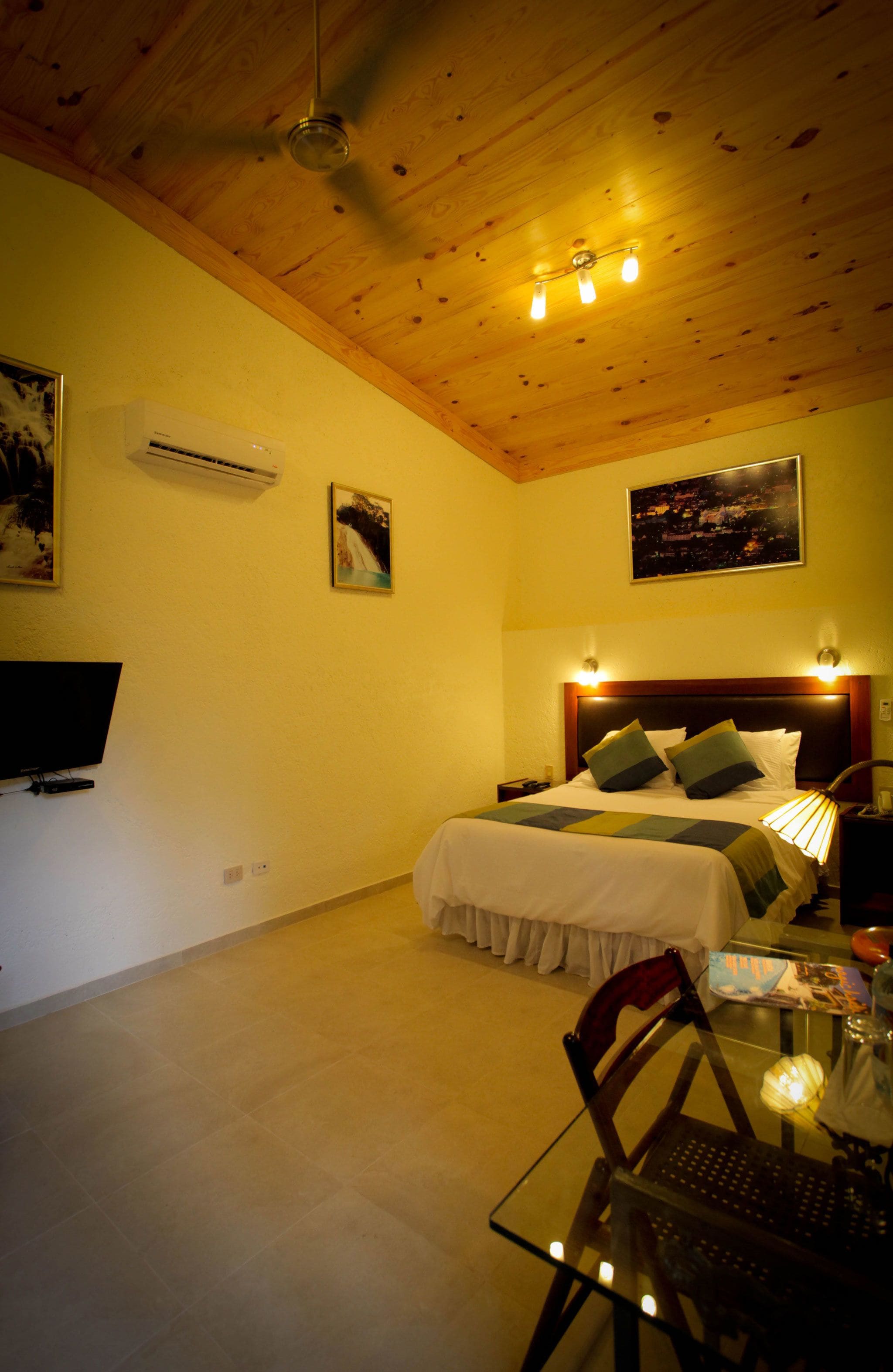 A bedroom with a wooden ceiling, a bed with pillows, a bedside table, a chair, a TV, and wall-mounted lights.