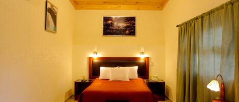 A hotel room with a wooden ceiling, a large bed with red bedding, two bedside tables with lamps, a nightstand with a phone, and a painting on the wall.