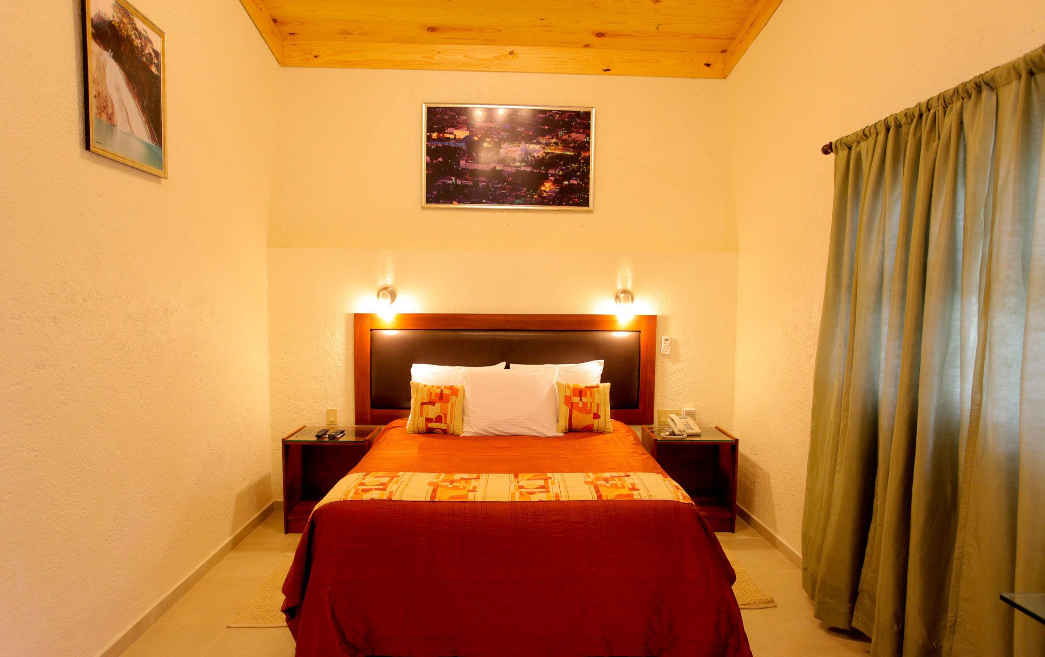 A hotel room with a large bed, two bedside tables with lamps, a nightstand with a telephone, a painting on the wall, and a view of a cityscape through the window.