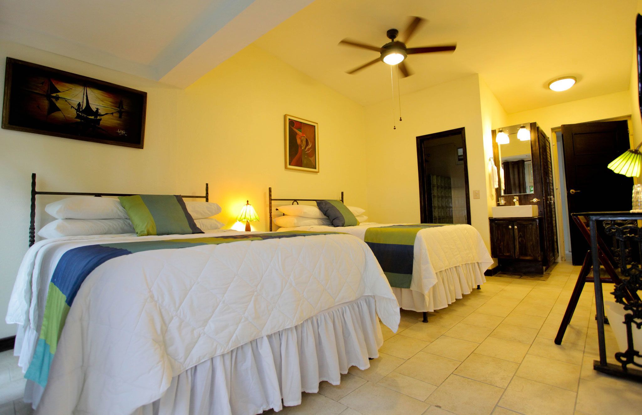A hotel room with two beds, a ceiling fan, a painting, a mirror, and a bathroom visible through an open door.
