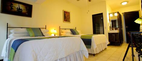 Habitación de hotel con dos camas, un ventilador de techo, un cuadro, un espejo y un baño visible a través de una puerta abierta.