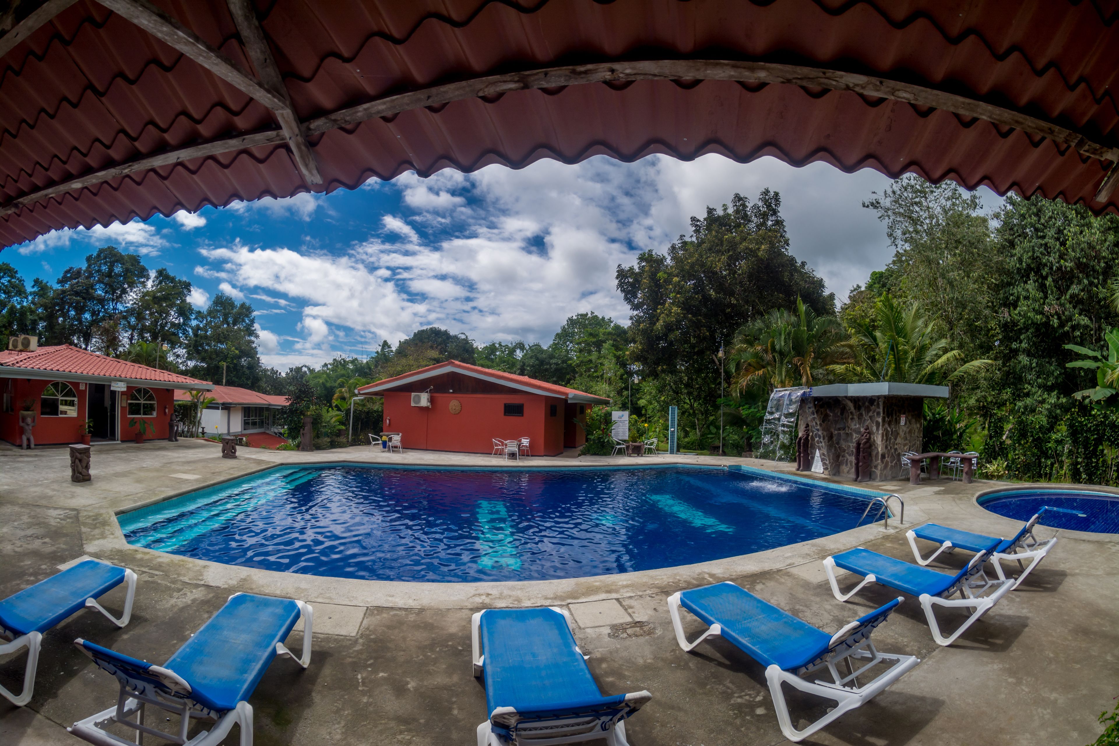 A swimming pool with blue water, surrounded by lounge chairs and a covered area with a red roof.