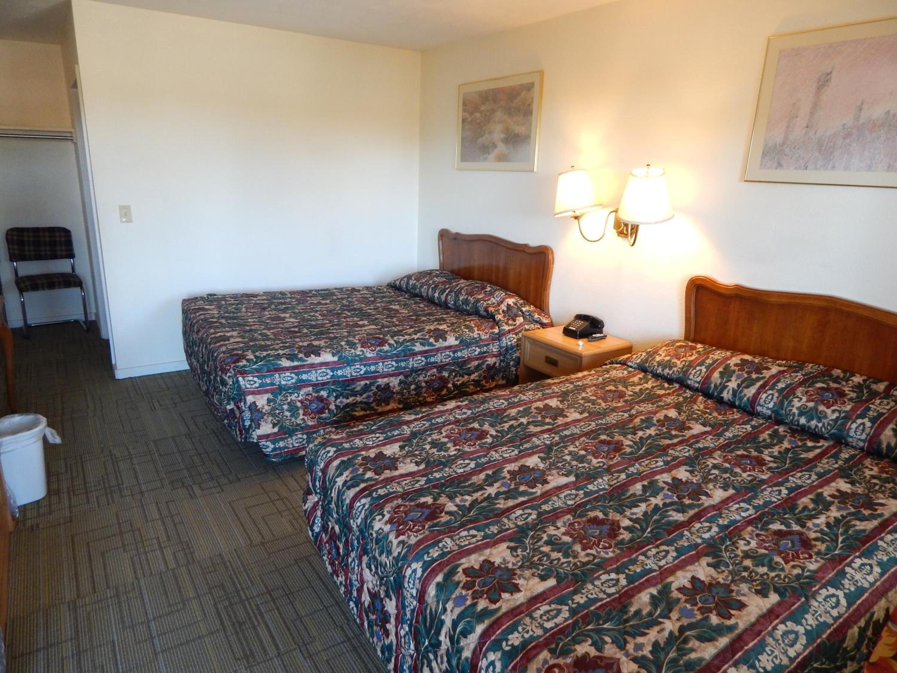 Quarto de hotel com duas camas, uma cadeira, mesa de cabeceira com abajur e um quadro na parede.