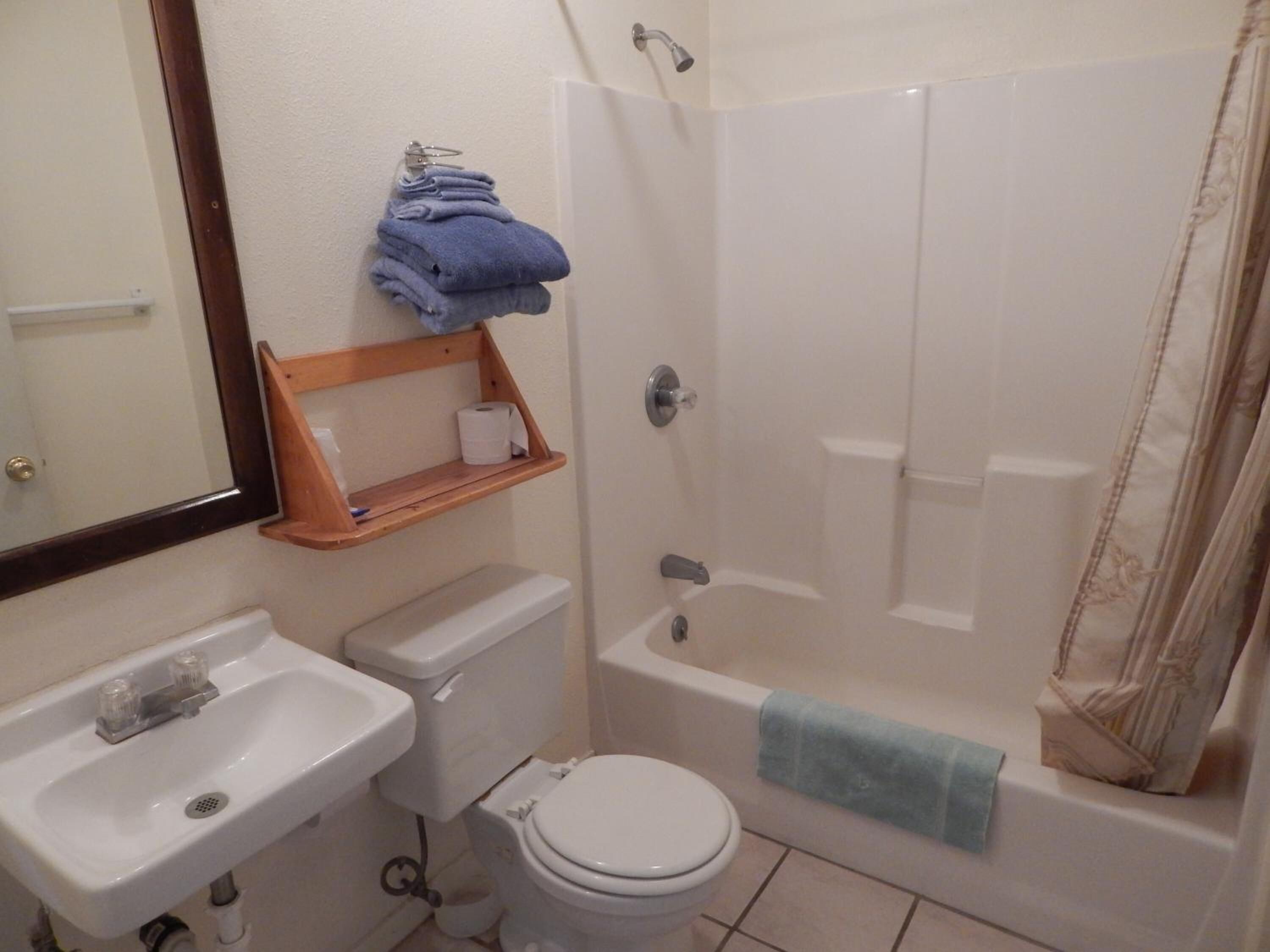 A bathroom with a sink, toilet, and bathtub. There is a shelf with towels and toilet paper.