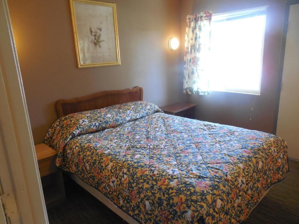 Ein Schlafzimmer mit einem blumigen Bettüberwurf, einem Nachttisch, einem gerahmten Bild an der Wand und einem Fenster mit Vorhängen.