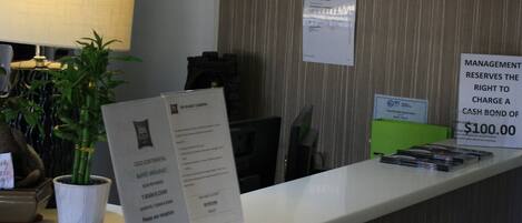Un mostrador de recepción de hotel con una pantalla de televisión mostrando un mensaje de agradecimiento, una planta en maceta, y carteles que indican los horarios del desayuno y el derecho de la administración a cobrar un depósito en efectivo.