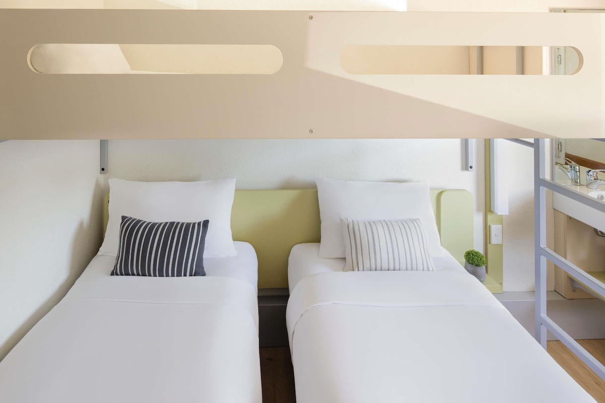 Habitación de hotel moderna con dos camas individuales, una planta pequeña en maceta y vistas al exterior.
