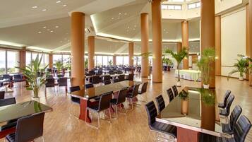 A spacious conference room with multiple tables and chairs, large windows, and several potted plants.