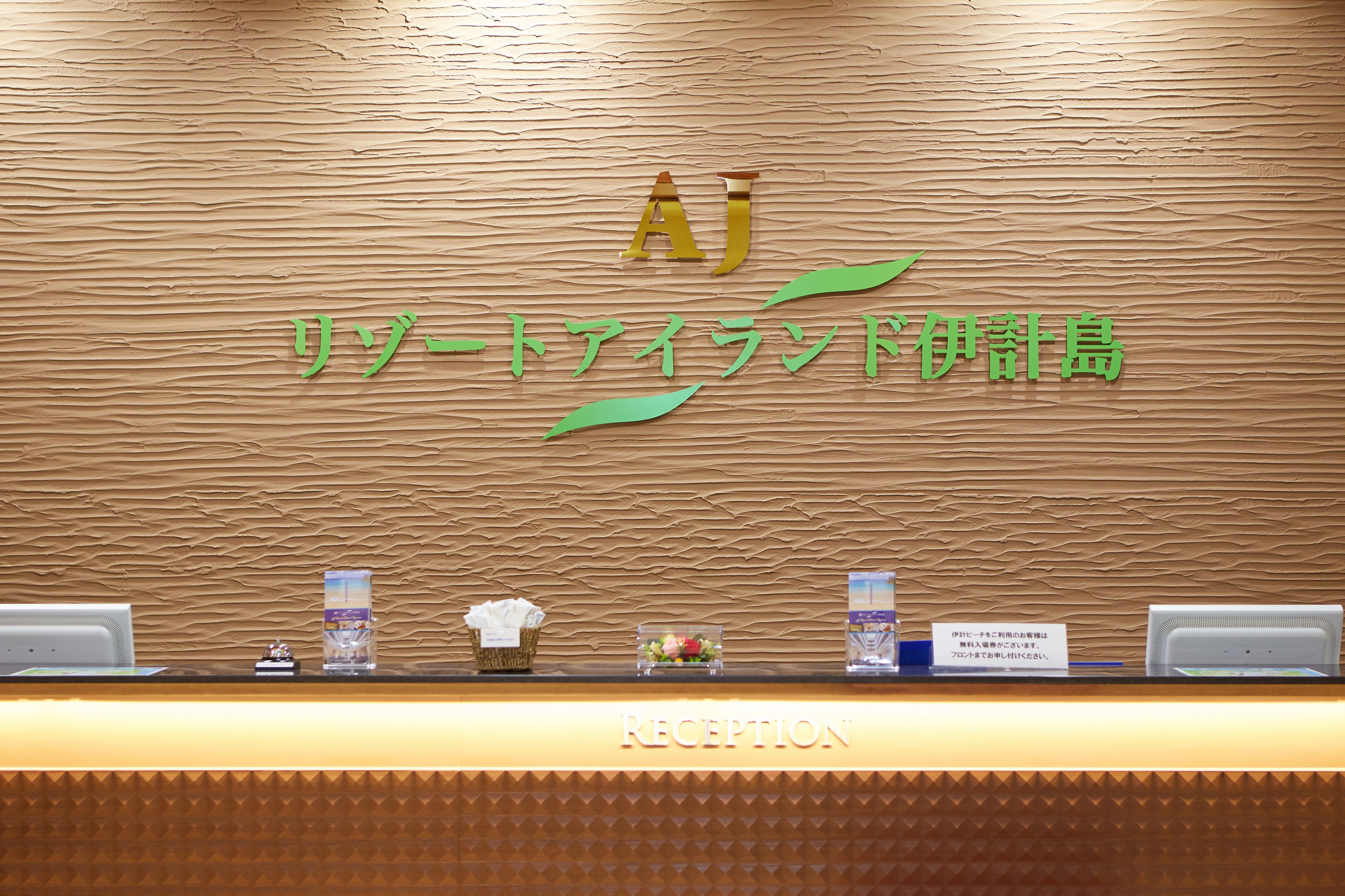 A reception desk with a wooden textured wall, a sign with the letters "AJ" and Japanese characters, and a reception area with a computer and a small basket with a tissue.