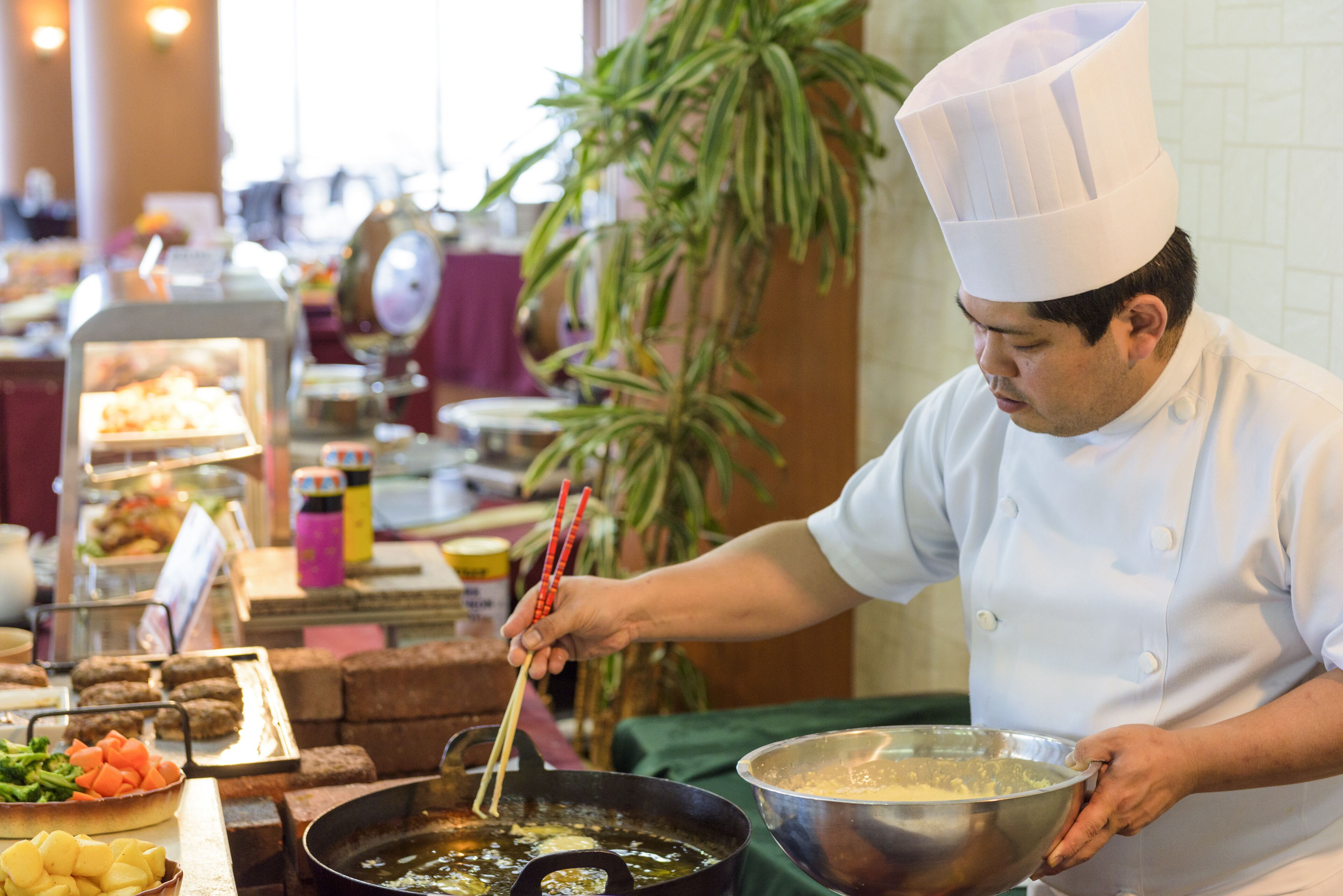 A chef in a white uniform and hat cooking in a large pan, with various food items and utensils visible.