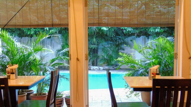 Un comedor interior con mesas y sillas, vista a una piscina y plantas en macetas.
