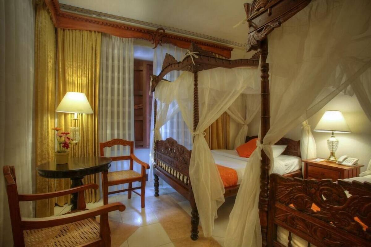 A four-poster bed with a canopy, a wooden chair, a small table with a lamp, and a bedside table with a lamp.