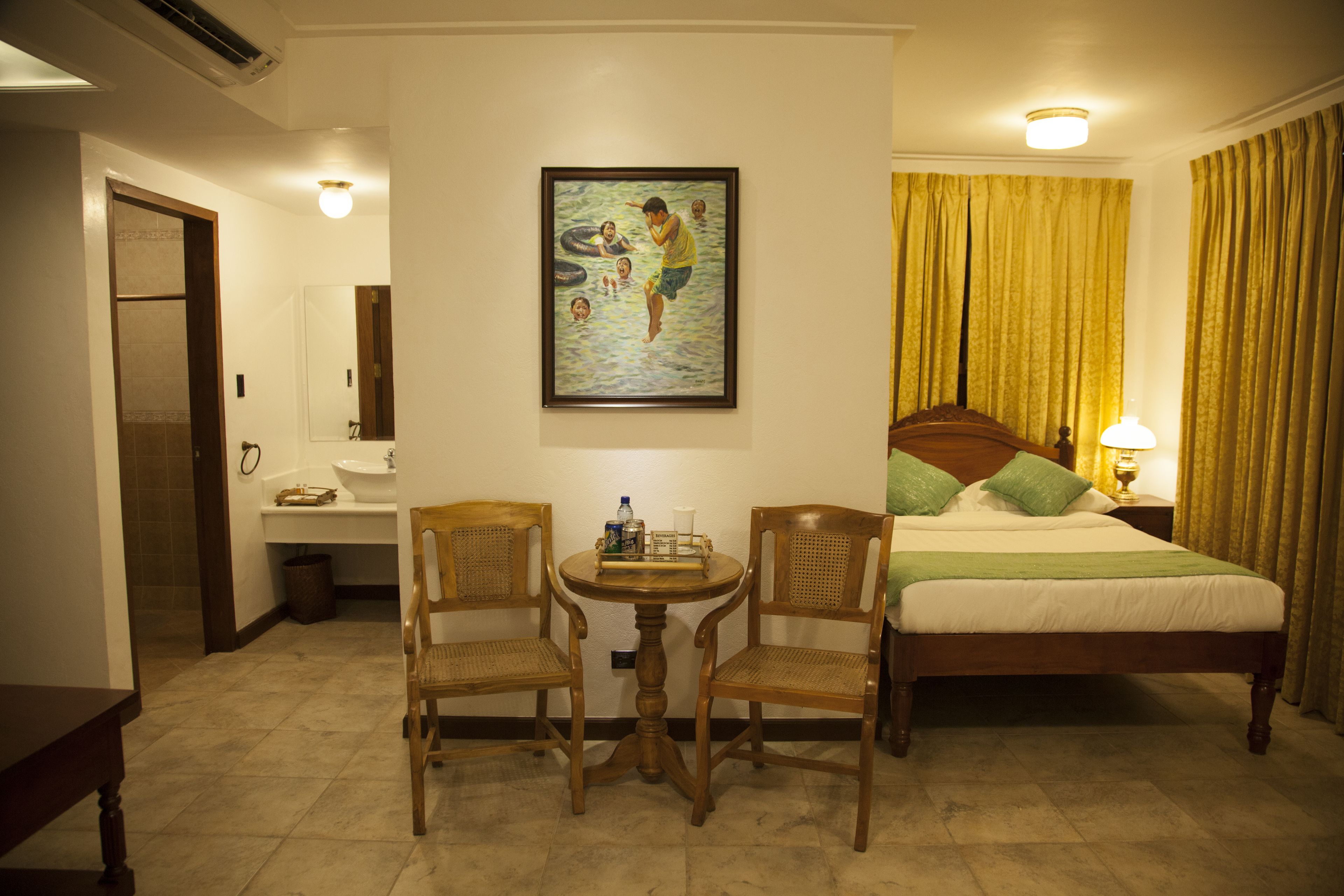Habitación de hotel con cama, dos sillas, una mesita, un cuadro, un lavamanos y un espejo.