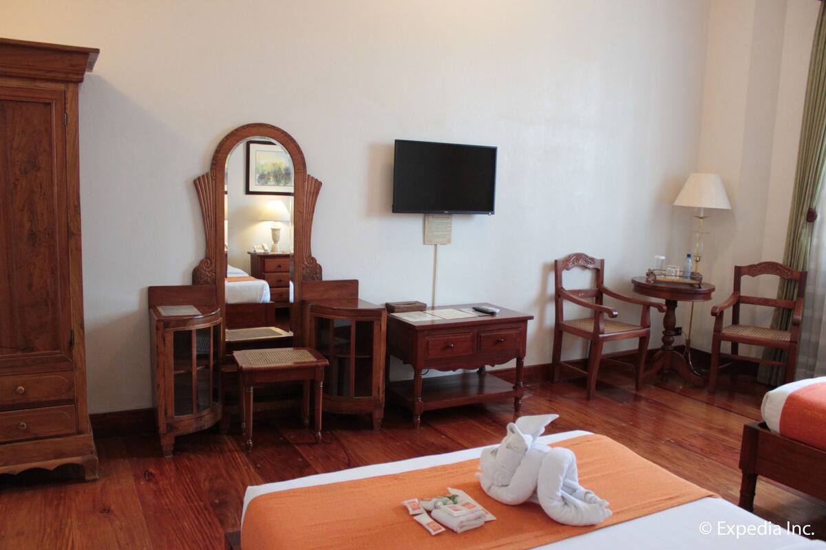 A hotel room with a wooden wardrobe, a desk with a chair, a bed with an orange bedspread, a television mounted on the wall, and a small table with a lamp.