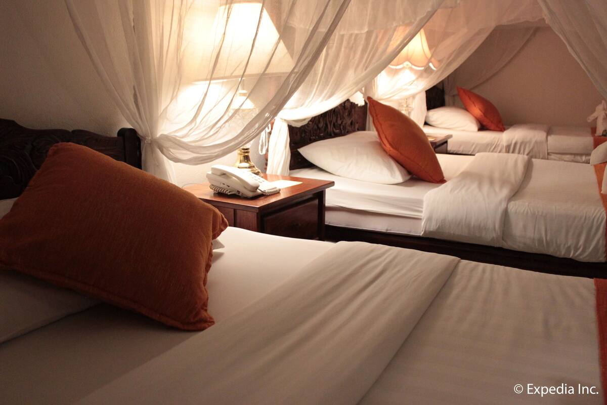 A hotel room with two beds, each with white bedding and orange pillows. There is a bedside table with a lamp and a telephone.