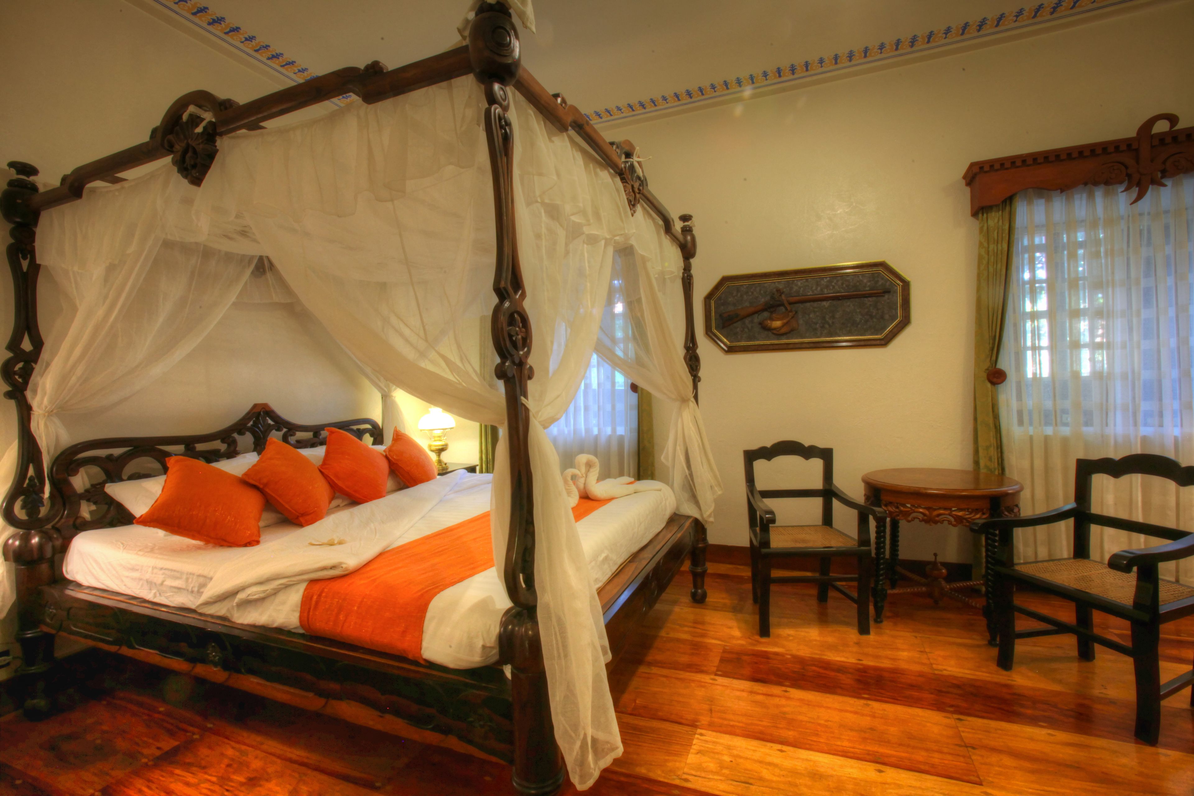A four-poster canopy bed with orange pillows, a wooden floor, two chairs, and a small table.
