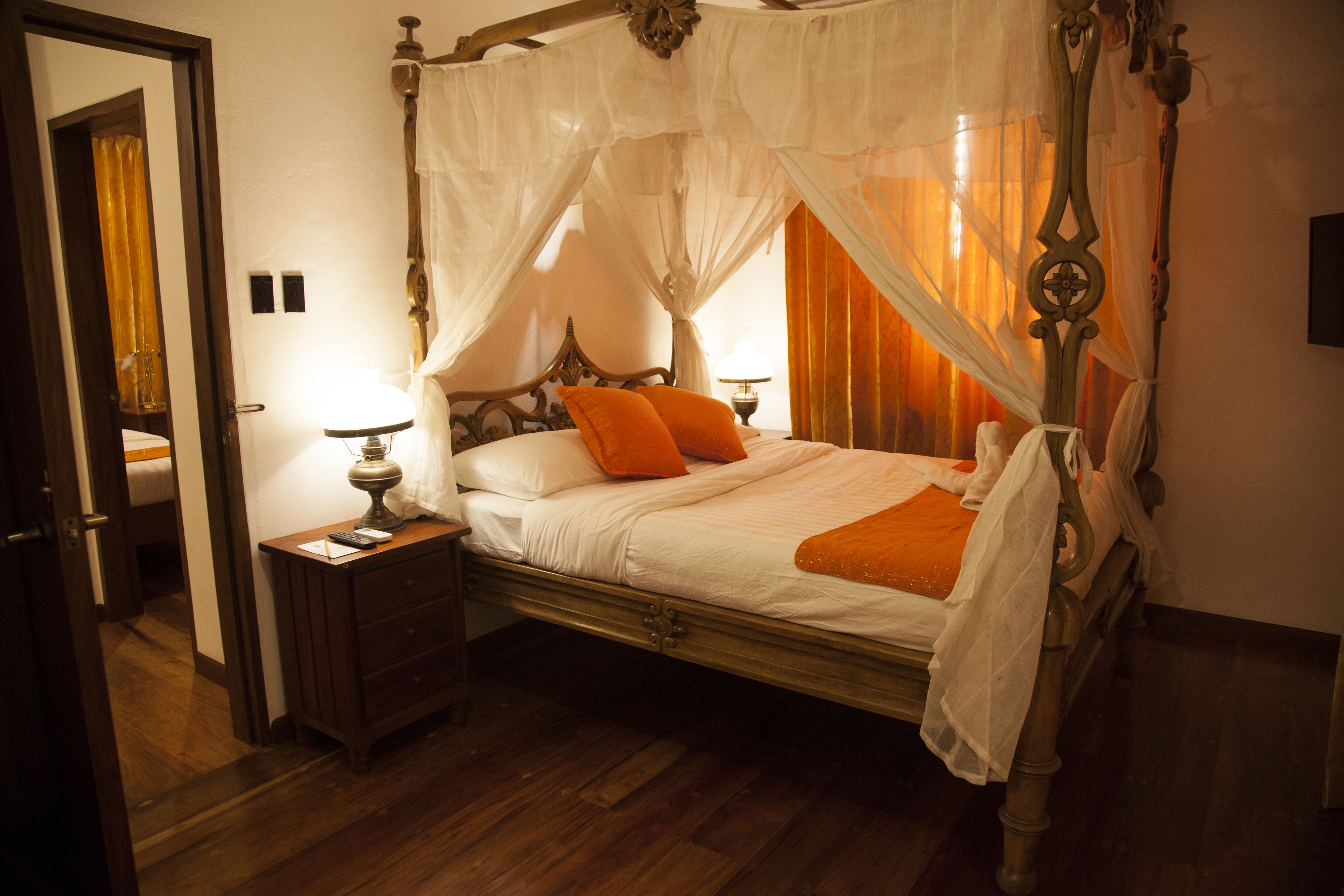 Una cama con dosel y cortinas, ropa de cama blanca con detalles naranja, una mesita de noche de madera con lámpara y piso de madera.