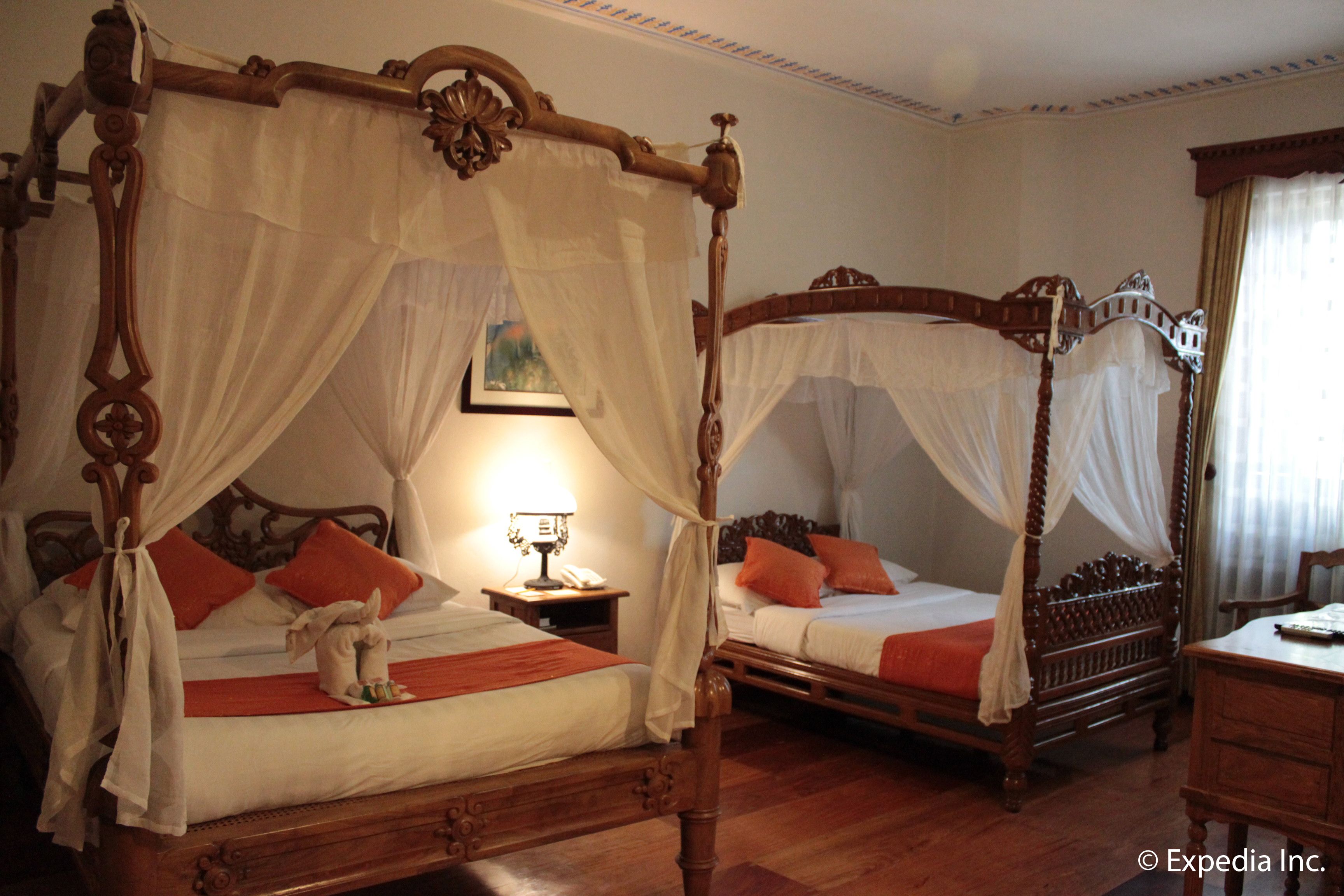 A room with two wooden four-poster beds, each with white bedspreads and orange pillows. There is a nightstand with a lamp and a framed picture on the wall.