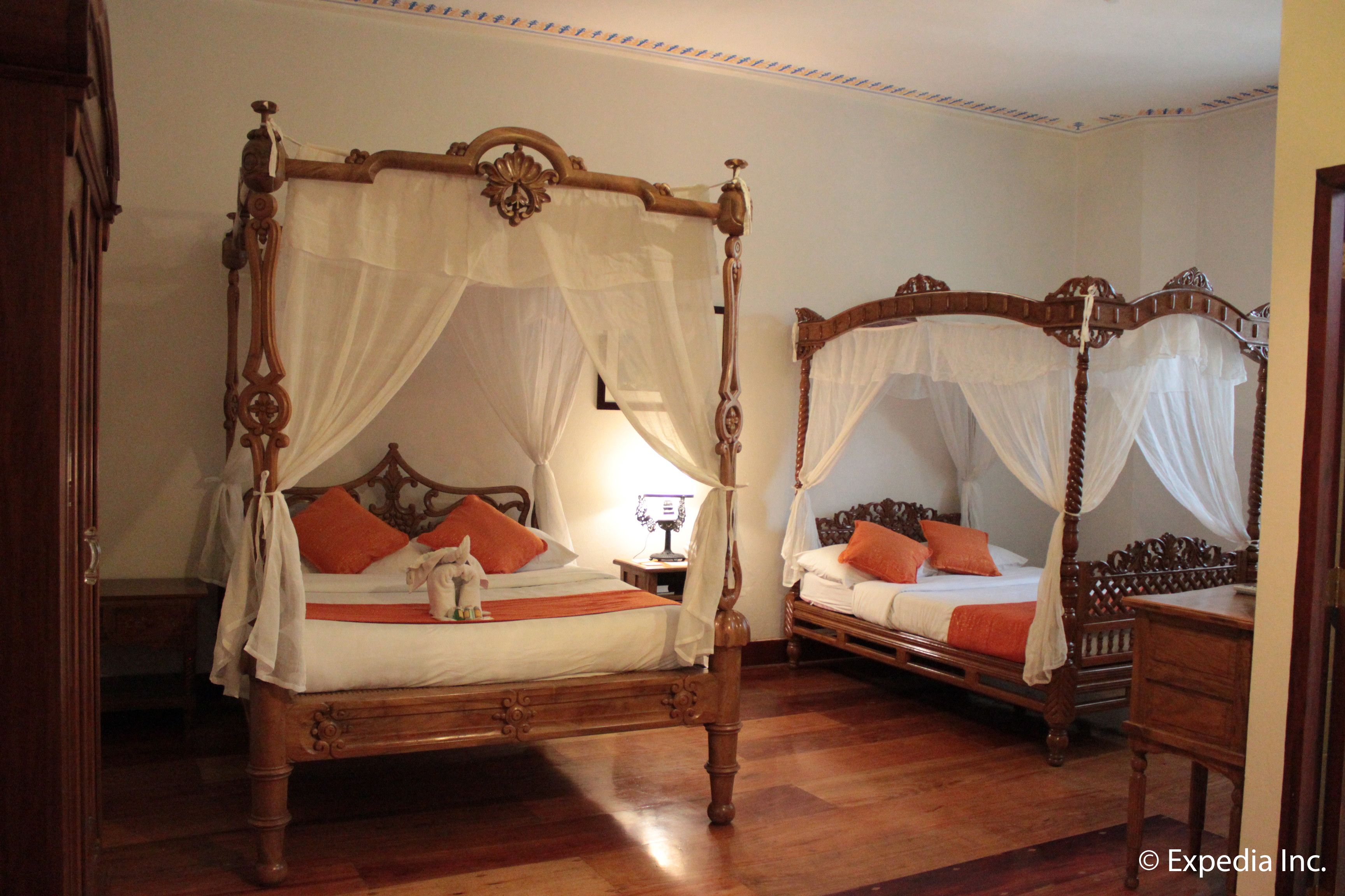 A room with two four-poster beds with white canopies, wooden floors, and a nightstand with a lamp.