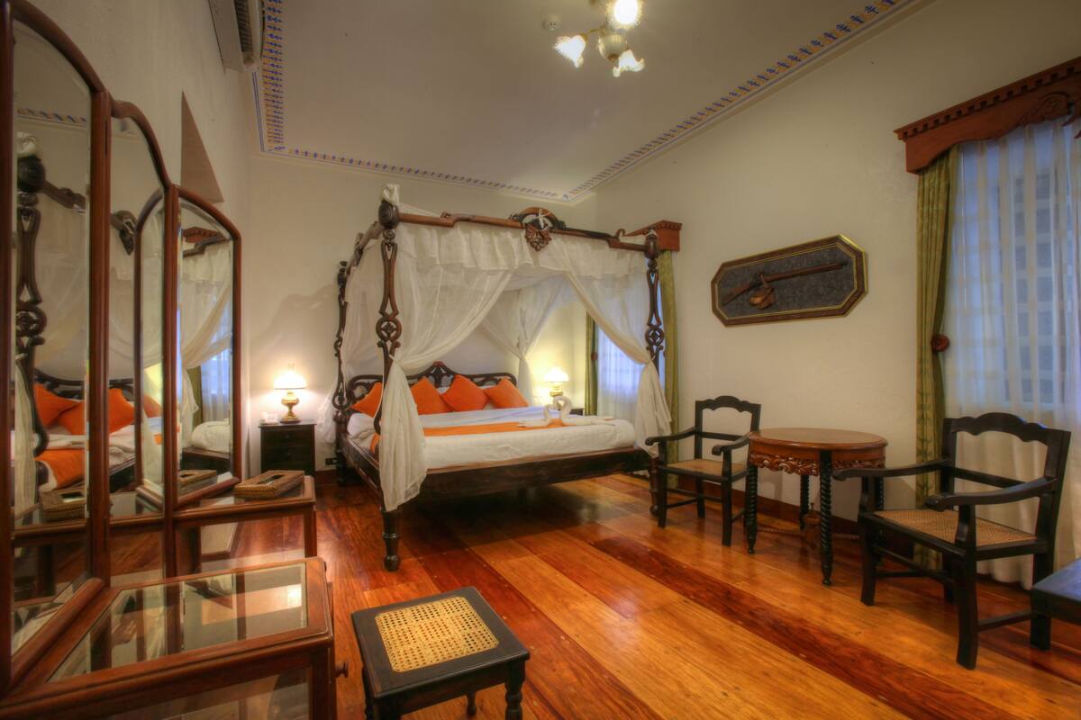 A four-poster bed with a canopy, a sitting area with a round table and chairs, and a mirror with a wooden frame.