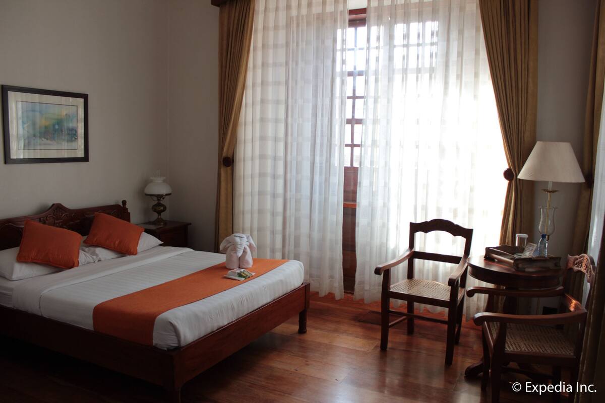 A hotel room with a wooden bed, two pillows, a bedside table with a lamp, a chair, and a window with sheer curtains.