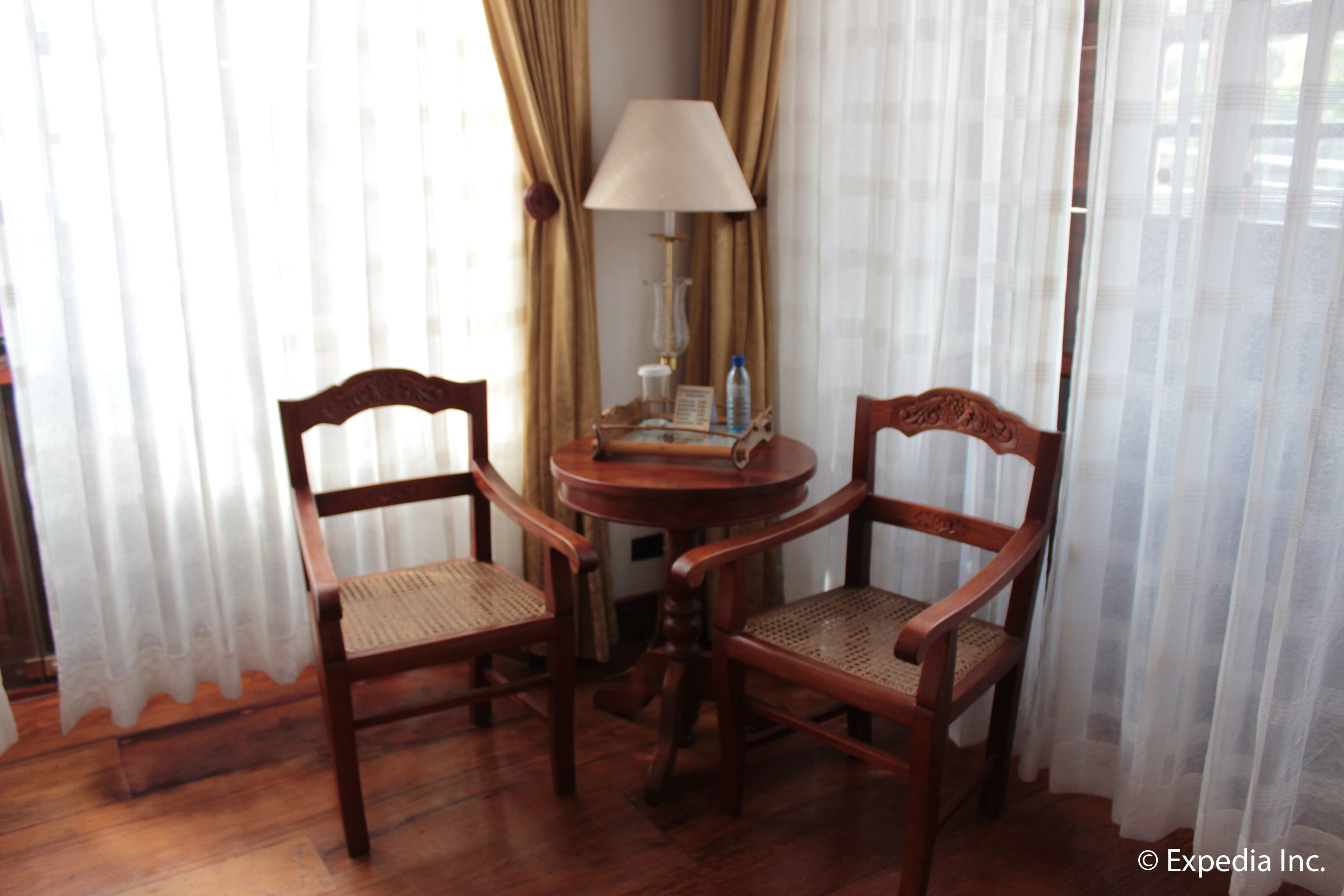 Una habitación con dos sillas de madera, una mesita redonda pequeña, una lámpara y una botella.