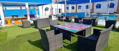 An outdoor seating area with wicker furniture and a patterned tablecloth, surrounded by a pool and a building with a blue roof.