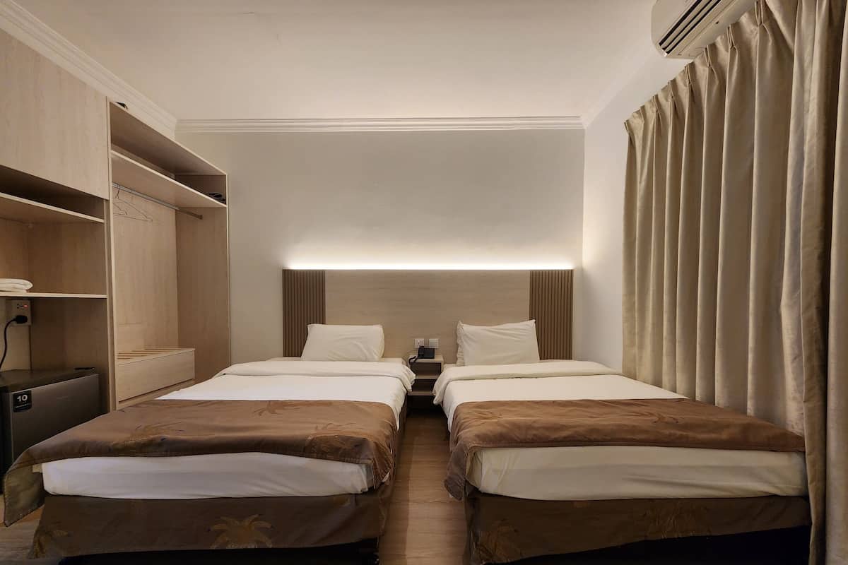 Habitación de hotel con dos camas, una mesita de noche con lámpara, un armario y un ventanal grande con cortinas.