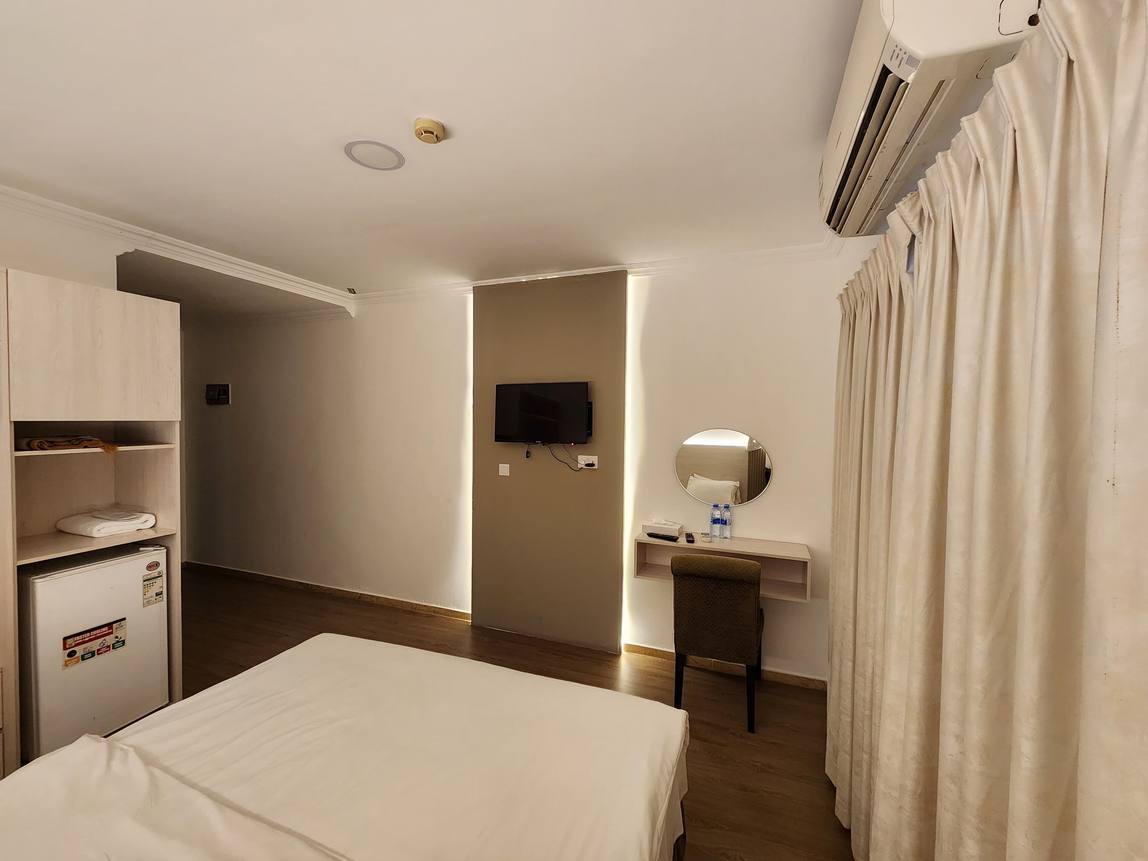 A hotel room with a bed, a desk with a mirror, a television, and an air conditioning unit.