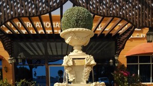 Una fuente de varios niveles con intrincados adornos y una ornamentación esférica en la parte superior, ubicada en una zona ajardinada con plantas en macetas y un edificio al fondo.