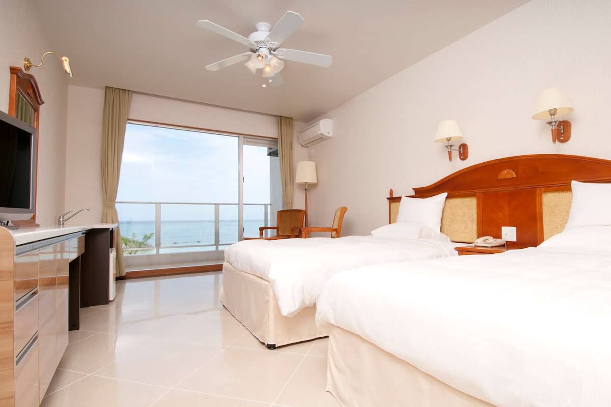 Habitación de hotel con dos camas, un escritorio, un televisor, un ventilador de techo y vistas al mar.