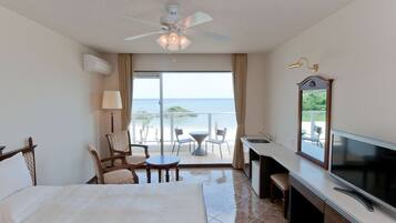 A hotel room with a bed, a desk, a TV, a balcony with a view of the sea, and a ceiling fan.