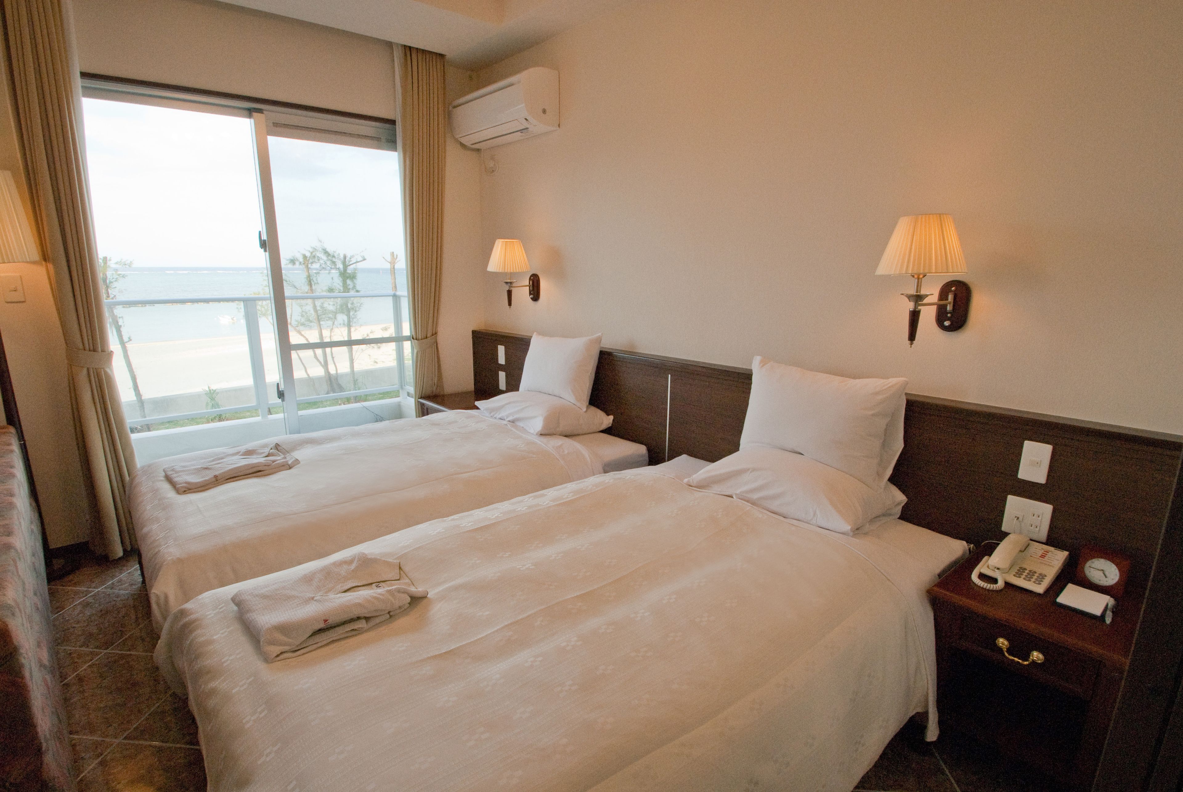Habitación de hotel con dos camas, una ventana con vista a la playa, una mesita de noche con teléfono y reloj, y un equipo de aire acondicionado en la pared.