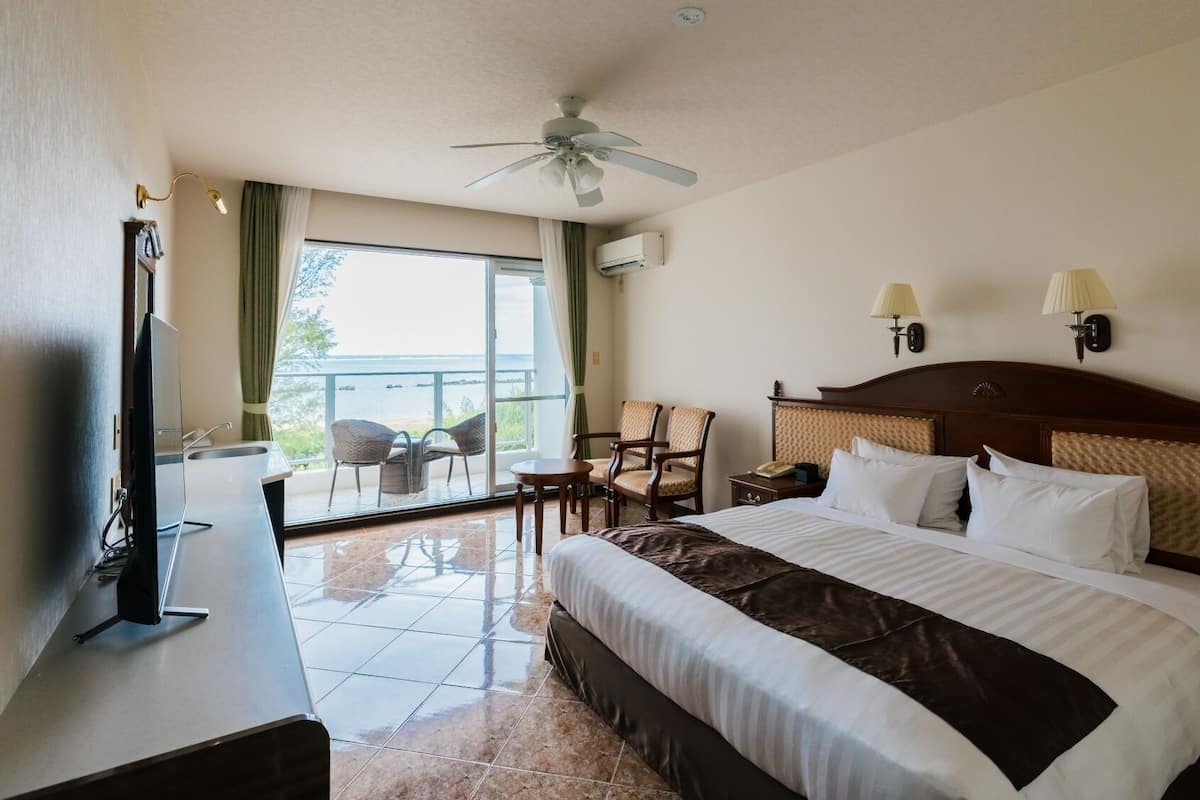 Una habitación de hotel con una cama grande, televisión, balcón con sillas y ventilador de techo.