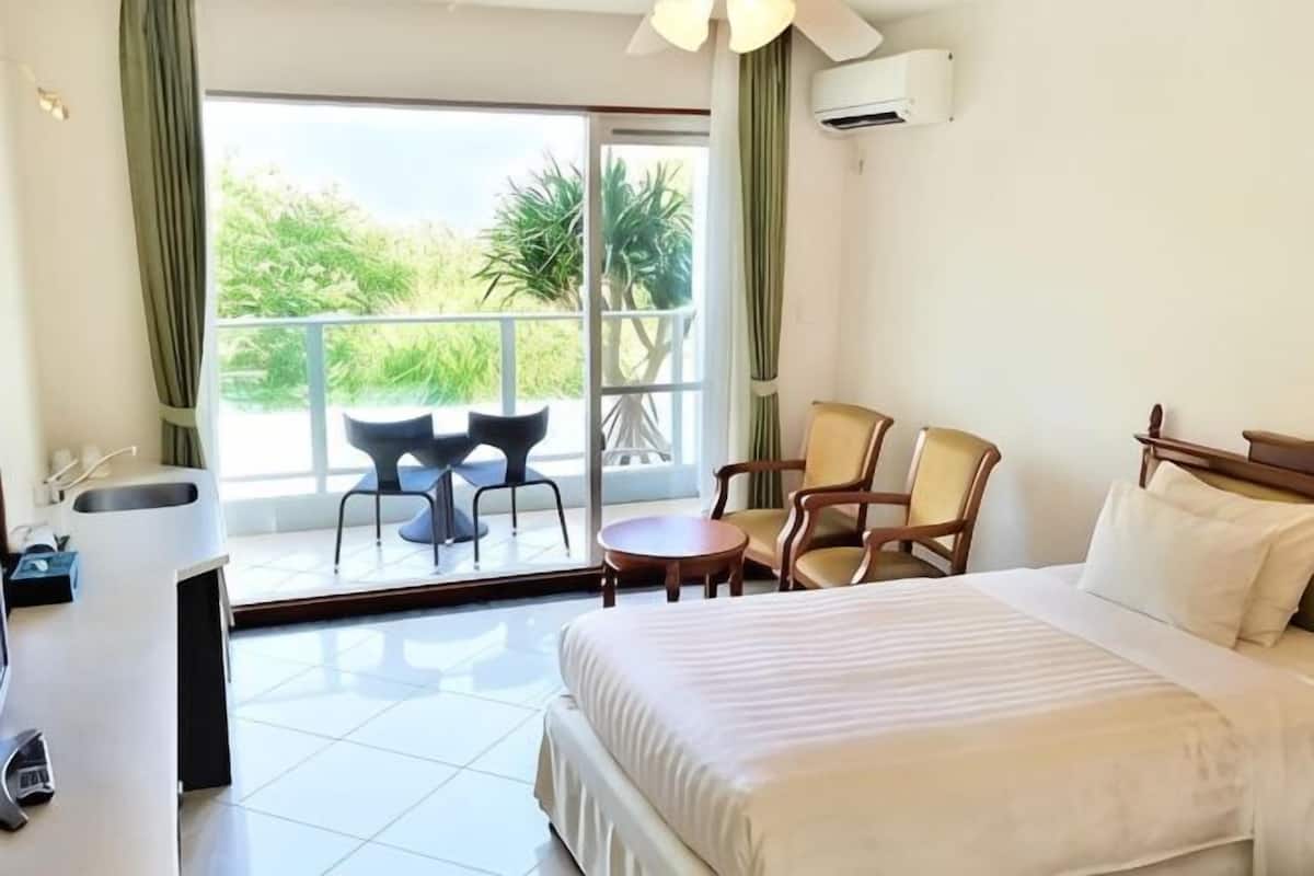Habitación de hotel con cama, una mesita, dos sillas, un lavabo y un balcón con vistas a la vegetación.