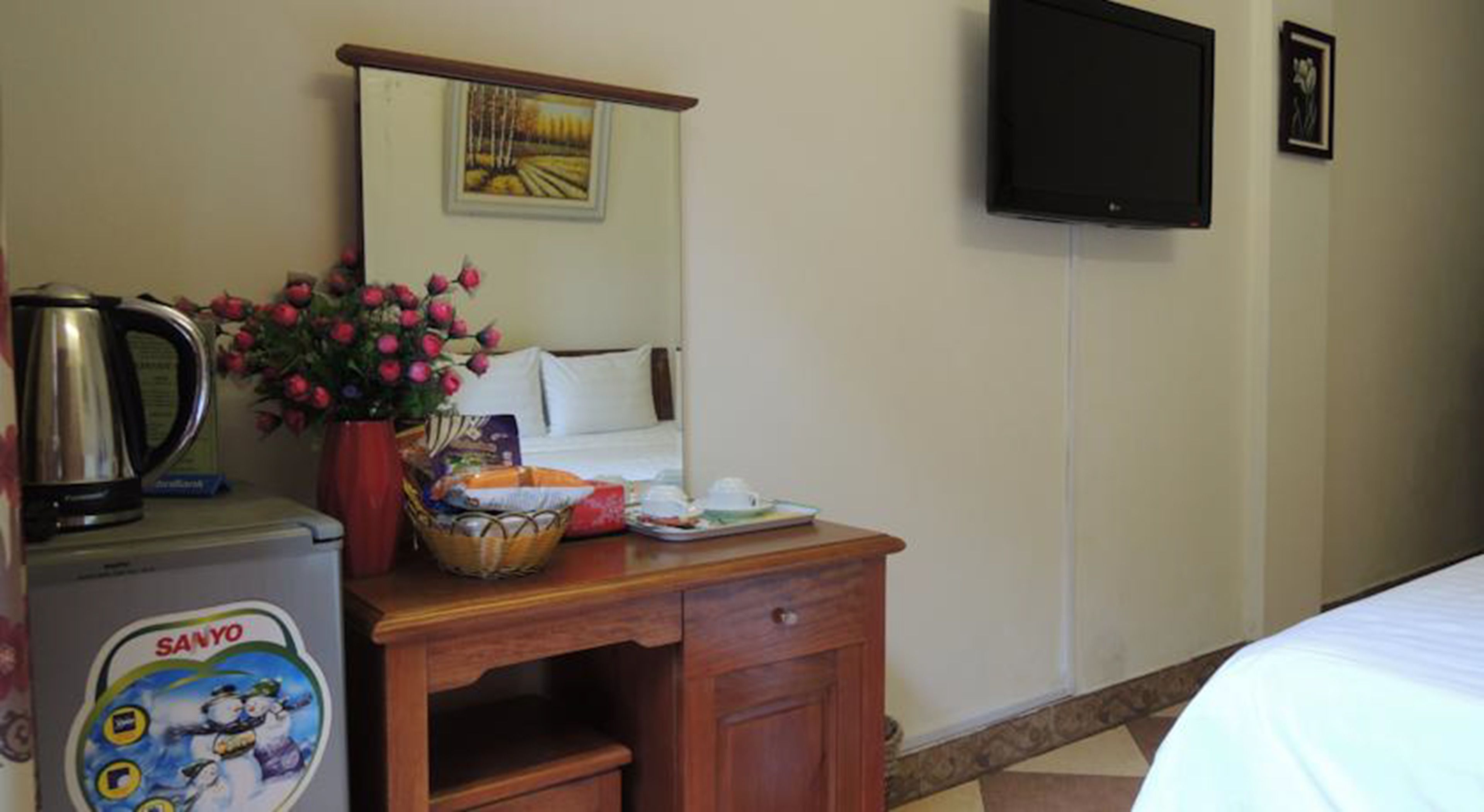 Quarto de hotel com uma secretária de madeira, chaleira, cesta de petiscos, vaso de flores, cama, televisão e um quadro na parede.