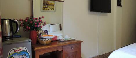 Habitación de hotel con escritorio de madera, tetera, cesta de aperitivos, florero, cama, televisor y un cuadro en la pared.