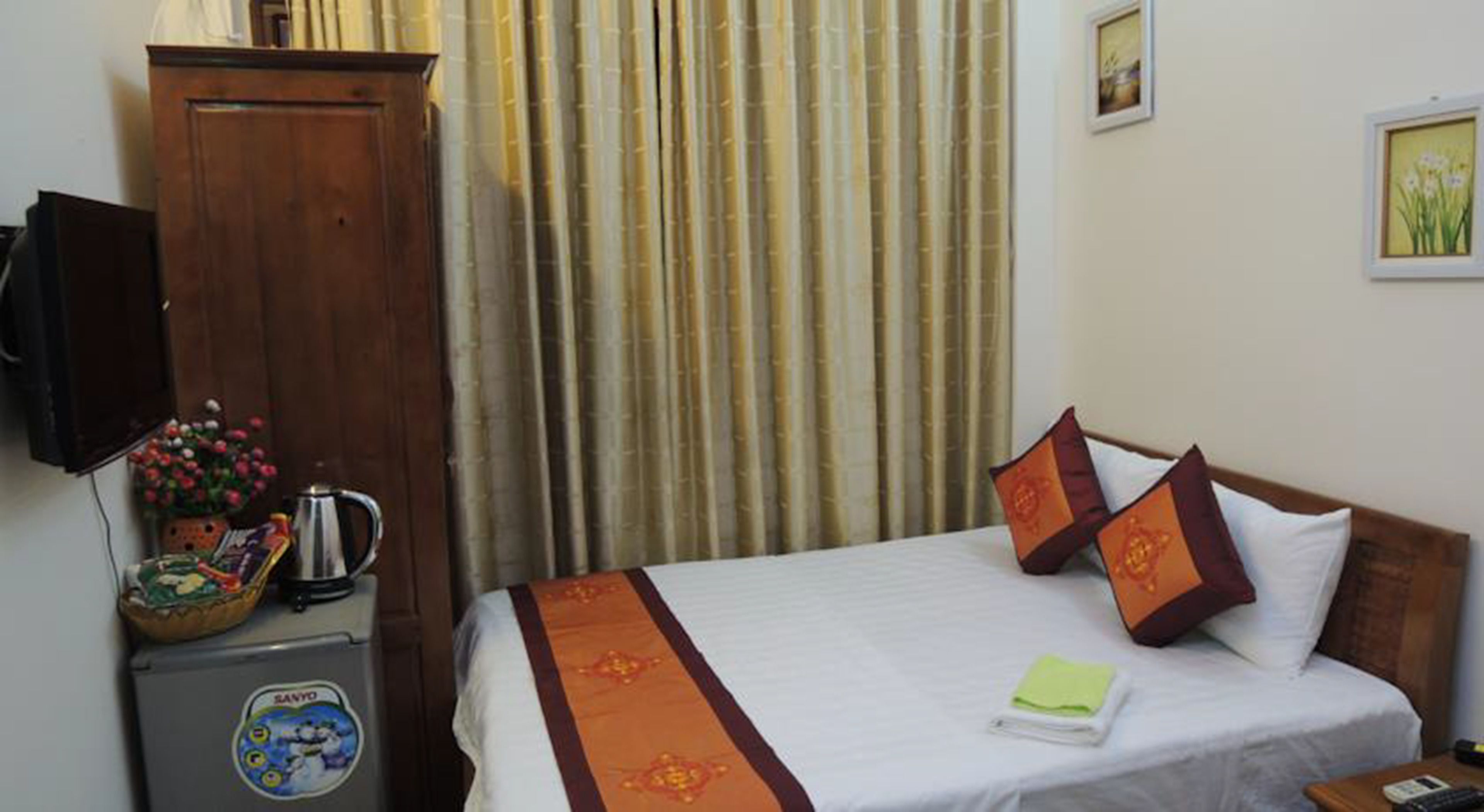A hotel room with a bed, a wooden wardrobe, a small table with a kettle and flowers, and a refrigerator with a sticker.
