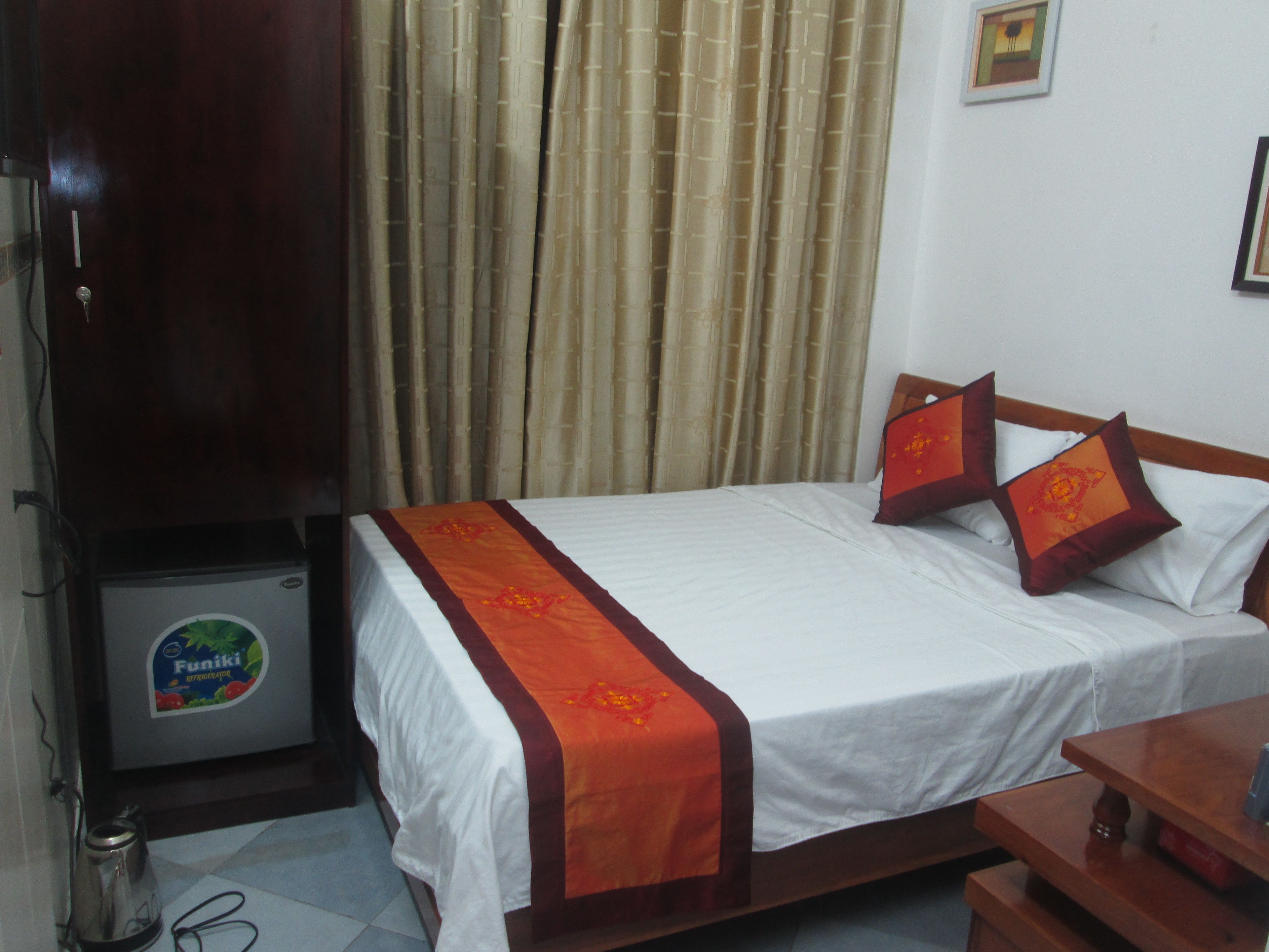 A hotel room with a bed, two pillows, a wooden nightstand, a small table, a kettle, and a refrigerator.