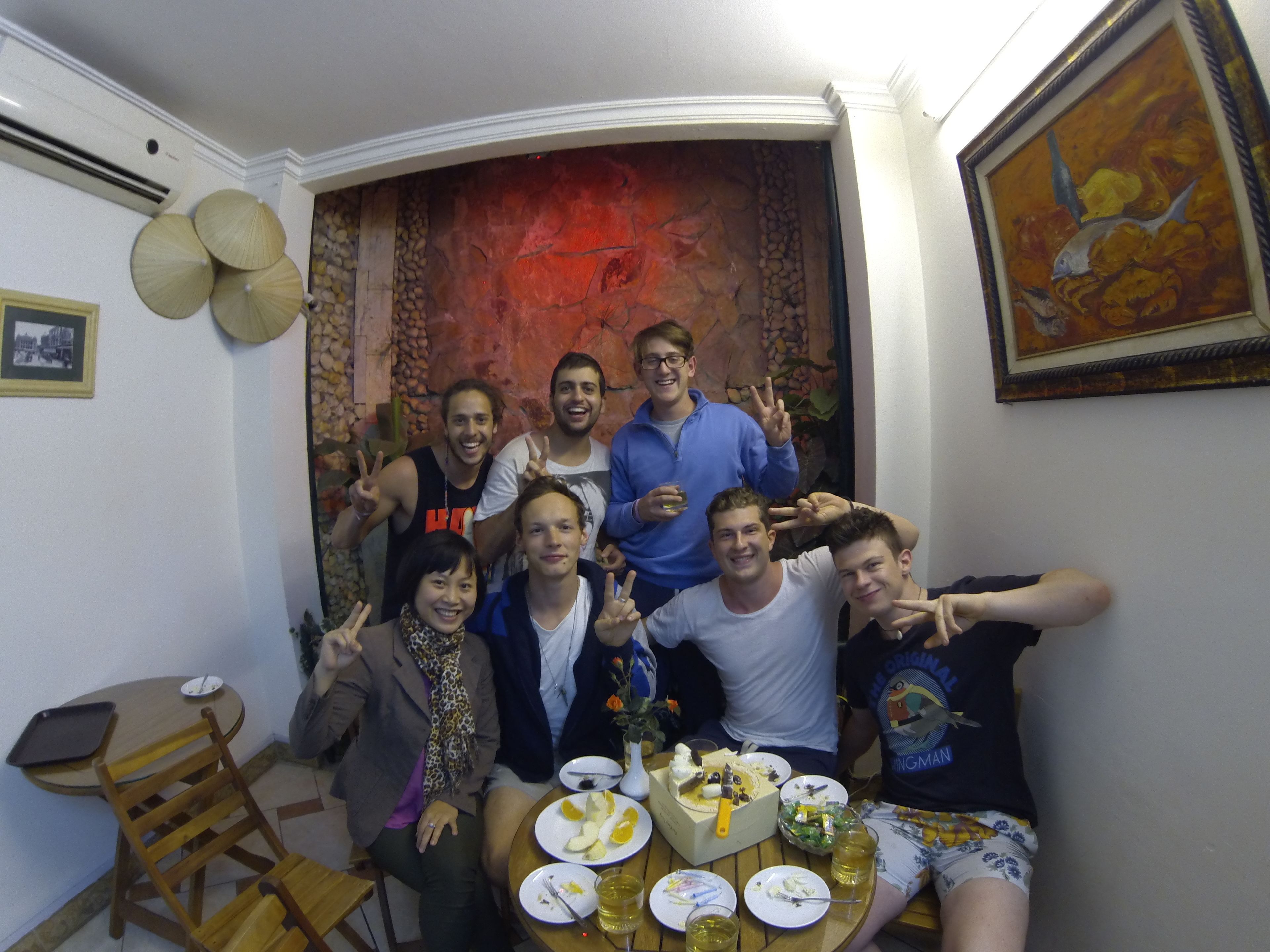 A group of people around a dining table with food and drinks, a painting on the wall, and a small table with a tray.