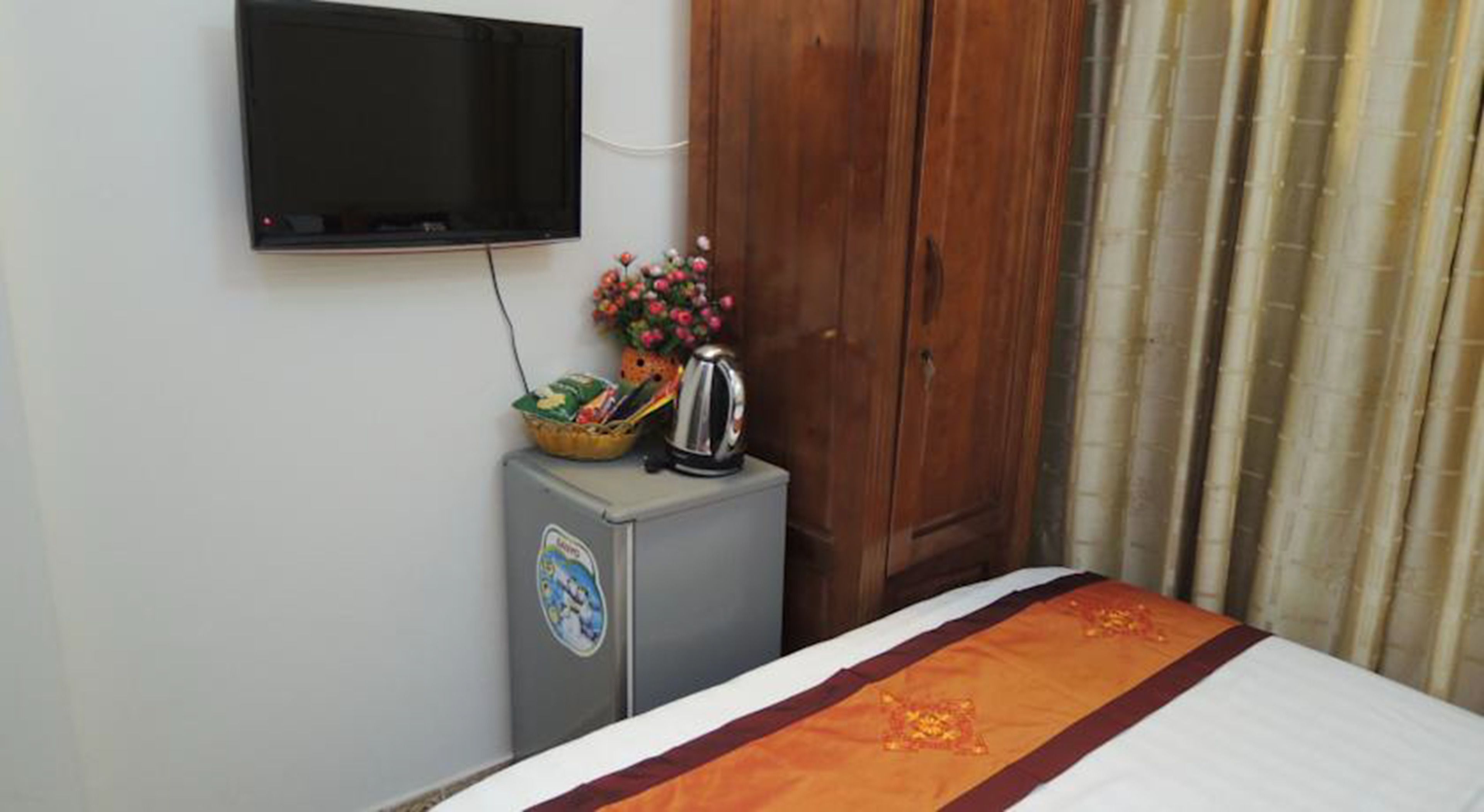 A hotel room with a bed, a TV mounted on the wall, a small cabinet with a kettle and snacks, and a wooden wardrobe.