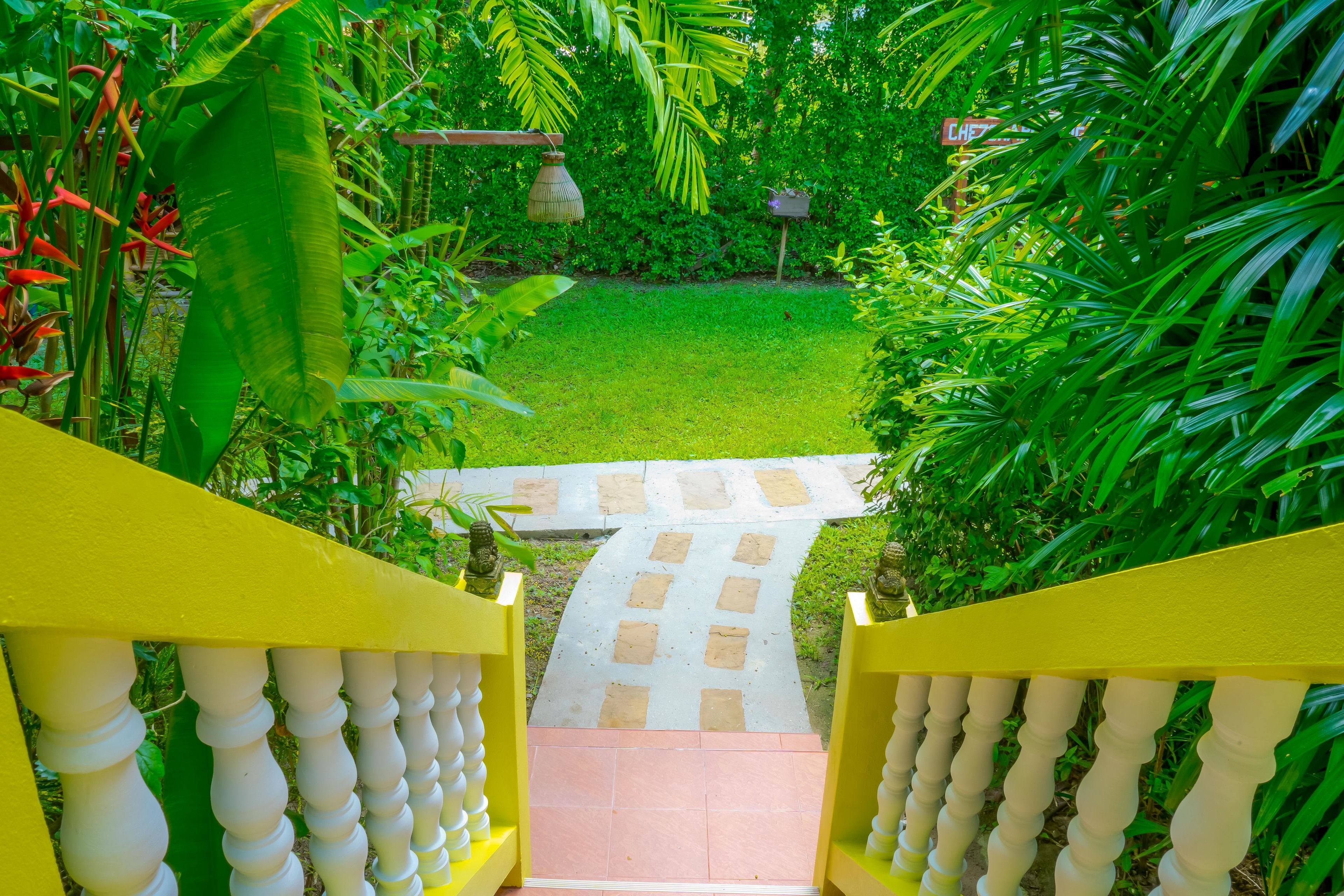 Caminho no jardim que leva à entrada de um edifício, com guarda-corrimão amarelo e chão de azulejos.