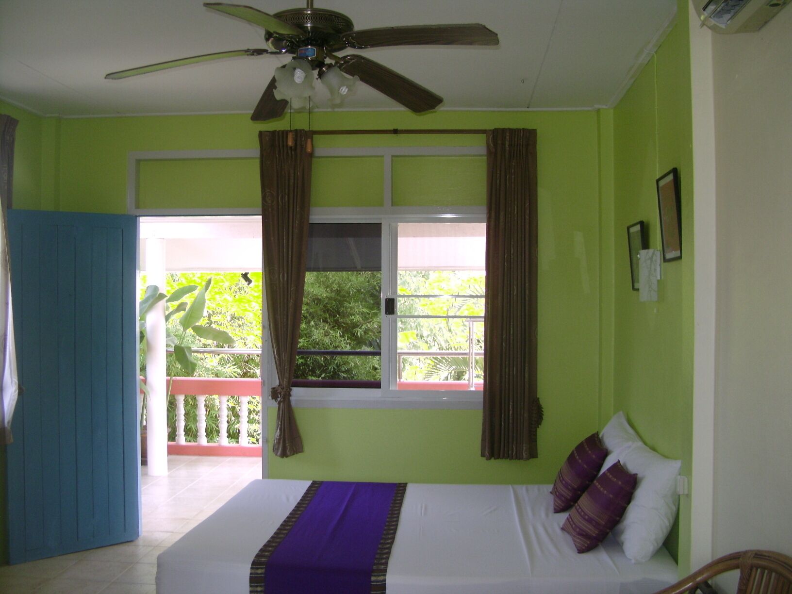 A room with a bed, a ceiling fan, a window with curtains, and a view of greenery outside.